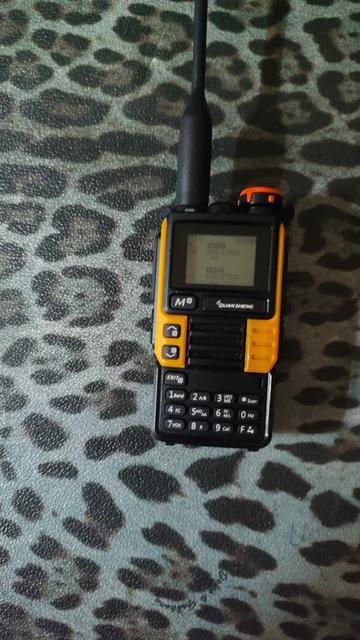 The image shows a handheld two-way radio or walkie-talkie device made by Quansheng. It has a black and orange/yellow color scheme. The device features a small LCD display screen at the top, with a long black antenna extending from the top. Below the screen are various control buttons including a numeric keypad (0-9) and function buttons. The radio appears to be sitting on what looks like a textured surface with a leopard-print or spotted pattern in gray and black colors. The device has a rugged, professional appearance typical of commercial or emergency communication radios.