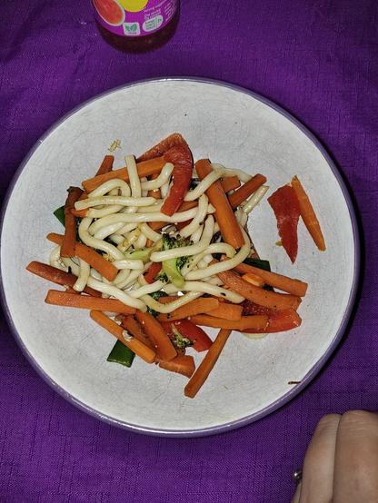 Young uns dinner: A (half eaten) bowl of stir fry veg with far noodles. Includes carrots, sugar snap peas, broccoli, red pepper and tofu. Set on a deep purple tablecloth.