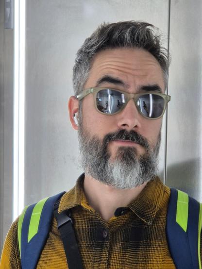 Selfie en el ascensor. Llevo una camisa de cuadros color mostaza y negro. La mochila para ir a entrenar y gafas de sol.
