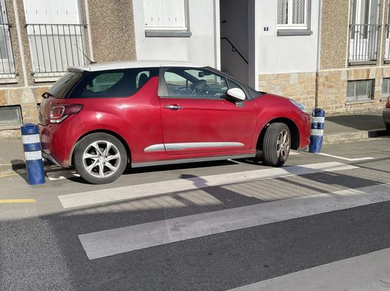 Voiture rouge stationnée sur un passage piéton pourtant encadré par 2 potelets bleus, la voiture touche quasiment les 2