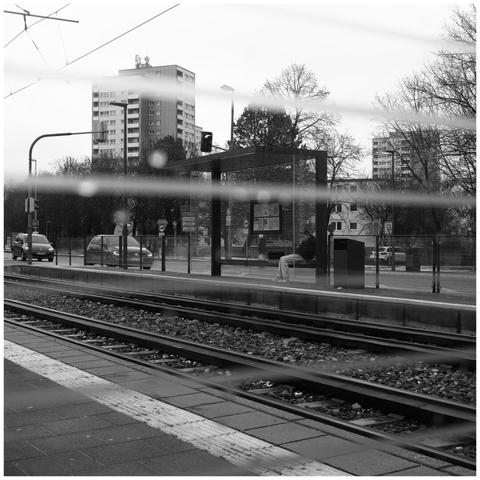 A person waiting at the opposite waiting area of a tram station. Photographed through the glass of the waiting shelter which has some lines printed on it.
