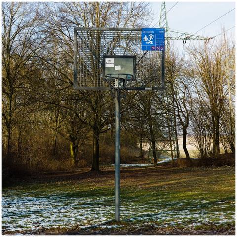 An all-metal outdoor basketball hoop in a park. A bit of snow lies on the ground.