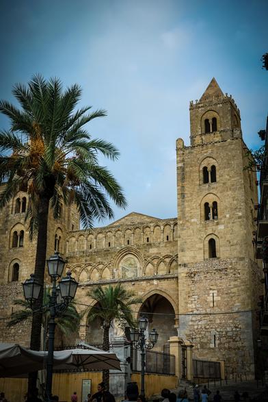 The image features a grand historical building, likely a cathedral, with two prominent towers. The architecture is characterized by stone construction, arched windows, and intricate stonework. The building's facade is adorned with a series of arches and a central rose window, typical of Gothic architecture. The towers are tall and feature pointed roofs, with arched openings and small windows. In the foreground, a tall palm tree with green fronds and yellow dates adds a touch of nature to the scene. Street lamps with multiple lanterns are visible, contributing to the historical ambiance. The sky is partly cloudy, providing a soft light to the scene. In the lower part of the image, people can be seen walking, and there are outdoor seating areas with umbrellas, suggesting a public space. The overall atmosphere is one of historical significance and cultural richness.

Provided by @altbot, generated privately and locally using Ovis2-8B