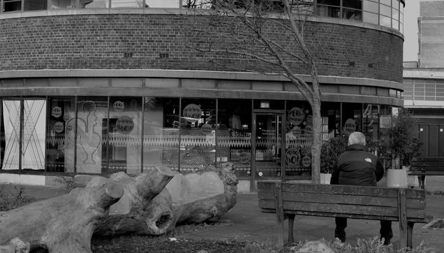 A person sits alone waiting on a bench, facing a windowed building with circular signs. A large tree log lies in the foreground, adding contrast to the monochrome, urban scene.

Eine Person sitzt einsam wartend auf einer Bank und blickt auf ein Gebaeude mit Fenstern und kreisfoermigen Schildern. Ein großer Baumstamm liegt im Vordergrund und bildet einen Kontrast zu der monochromen, staedtischen Szene.
