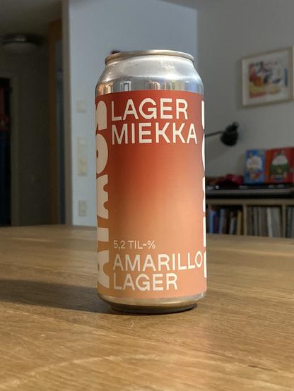 A can of Amarillo Lager beer resting on a wooden table. The can features a gradient design with the text "Lager Miekka" and "5,2 TIL-%" prominently displayed.