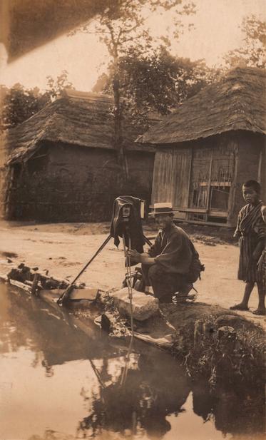 A man crouches at the edge of a river or creek next to an early 20th century camera. A boy stands to one side and a pair of houses with wooden walls and grass/thatch roofs can be seen in the background.