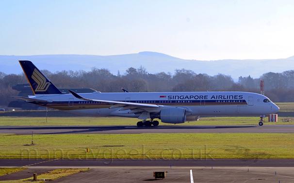 Side view of a white, twin engined jet airliner moving from left to right along a black runway.
The plane has a purple, orange, and gold stripe running along the body from just behind the cockpit, covering the passenger cabin windows, with the words "Singapore Airlines" on the upper forward fuselage in purple.
The tail is purple, with a large, golden, stylised flying bird.
The foreground grey apron and a black taxiway, separated from the runway by a wide strip of grass.
More grass in the background runs up to trees in the distance.
There is a large, green fire training rig in the background on the left of the frame.
Rolling hills in the far distance slowly vanish into bright but hazy sky.