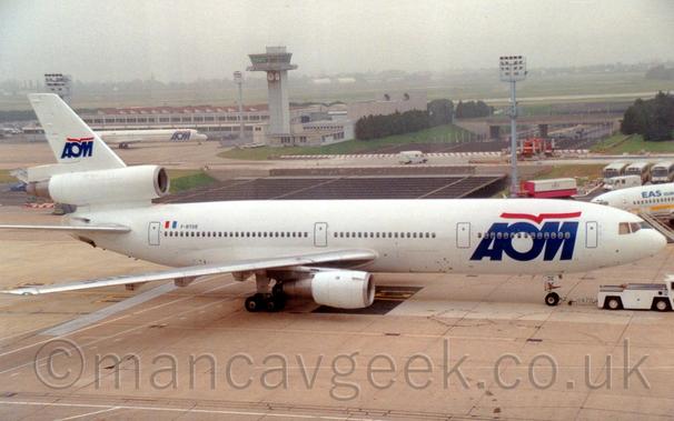 Side view of an almost all-white, 3 engined jet airliner being towed from left to right by a low-slung white tug attached to the nose-wheel.
There are large, blue, billboard-style "AOM" titles on the forward fuselage, with stylised red flying bird that resembles an almost straight line over the top, a logo repeated in a smaller form on the tail.
There is a smaller, white jet airliner with the same logo on it's forward fuselage in the background on the left, parked in front of a white building with multiple red slanted roofs.
Next to that building, towards the centre of the image is a squat, grey building with a tall grey tower poking up from corner, with a heavily glazed top level and several clock faces suspended underneath.
Grass leads off into the distance, slowly vanishing into murky grey haze.