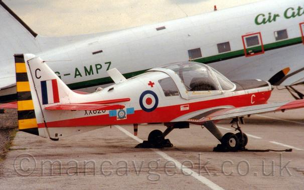 Side view of a white and red, single engined light aircraft parked facing to the right.
The top half of the body is white, the lower half is red, and the horizontal tail-plane and ends of the wings are also red.
There is a red, white, and blue roundel just aft of the over-sized, bulbous cockpit canopy, and a blue, whit, and red fin flash on the white tail.
The serial "XX620" is on the rear fuselage in black, partially obscured by the tail-plane.
 A larger, white, World War 2-era airliner is parked in the background, with a green stripe running along the body, and the registration "G-AMPZ" in green on the rear fuselage.