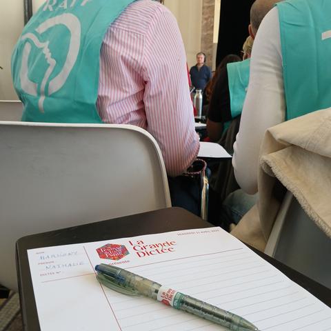 Une feuille de papier avec une entête La grande dictée, un stylo et devant des personnes avec une chasuble de la RATP
