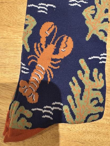 A close-up of a patterned sock featuring a large orange lobster and green seaweed against a navy background, with some white wave designs. The toe of the sock is a contrasting reddish color.