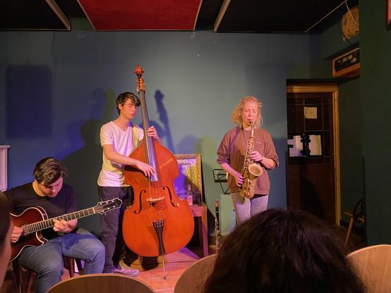 Three musicians playing on a small stage. From left to right they are playing guitar, double-bass and alto-saxophone.
