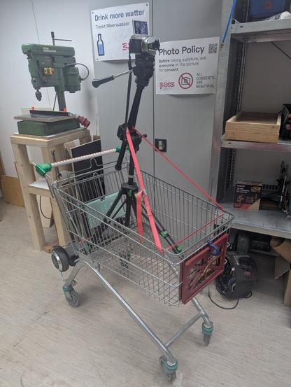 Ein Stativ samt Kamera in einem Einkaufswagen, welche mit einem roten Spanngurt an diesen befestigt ist. Im Hintergrund ist ein Teil vom Maschinenraum im Space zu sehen mit u.a. einer kleinen Standbohrmaschine links, einem Regal rechts und ein Schrank hinter dem vorhin genannten Konstrukt. Am Schrank hängen "Photo Policy" und "Drink more watter" Schilder vom 38C3.