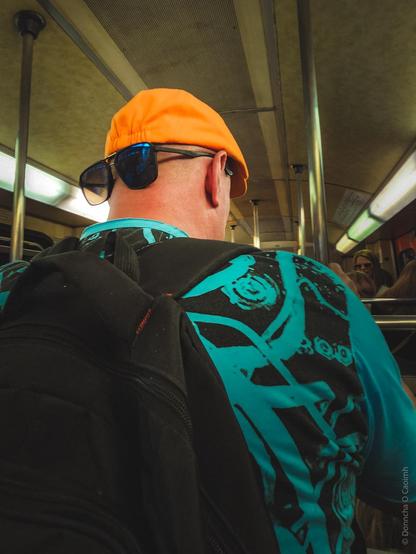 A man wearing an orange cap and sunglasses on the back of his head, dressed in a turquoise and black patterned shirt, with a black backpack, standing inside a metro train. The train's interior features metal poles, fluorescent lighting, and a beige ceiling.