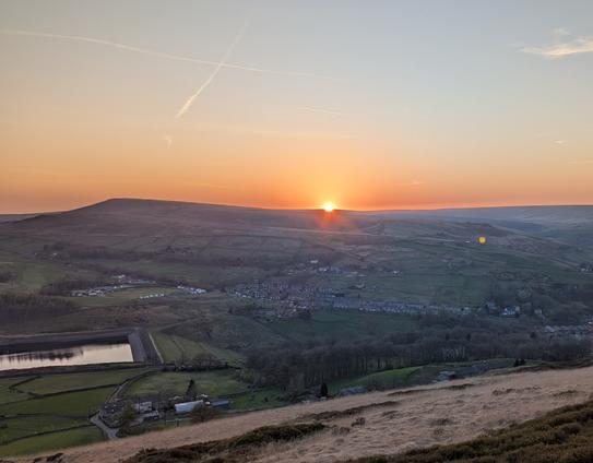 Sun half gone behind opposite hilltop across a moorland valley.  A reservoir, farms, and small town in the valley