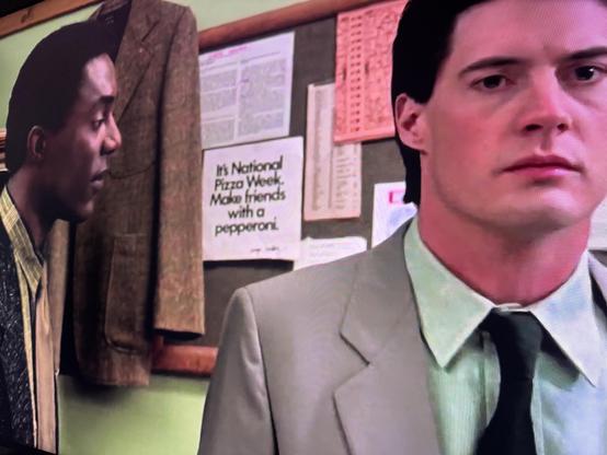 A scene featuring two men inside an office. One man in a gray suit looks serious, while the other, in a patterned jacket, appears to be engaged in conversation. Behind them is a board with a poster stating, "It's National Pizza Week.