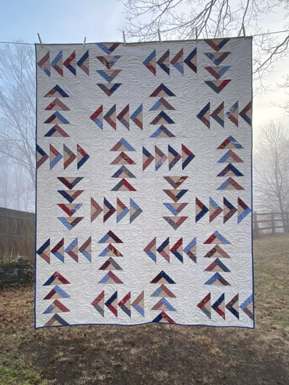 Quilt on a clothesline in earning morning fog of flying goose blocks in reds and blues with tan center triangles against a cream background.