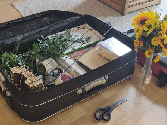 A luggage full of art items, decorations and fake plants. Next to it stands a bouquet of sunflowers and a pair of scissors