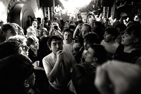 Black and white landscape orientated photo of a crowd in a rock concert. They are surrounding the singer who has come out into the audience and is singing into his microphone