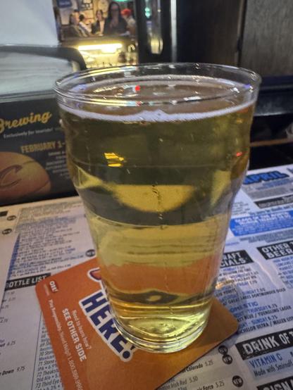 A pint glass filled with a golden beverage is placed on a coaster, set against a menu with various drink options. The background features people in a bar setting.