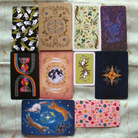Photo of the back image of the cards from the decks of the first photo. Again very different in style, colours, and size. Some feature plants, hands, hares, stars, and cats.