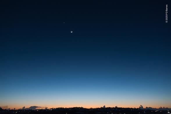 A tiny crescent moon and Venus sit high in the immense, deep blue vault of a dawn sky. Overhead the sky is almost black, slowly graduating through lighter and lighter blues almost to the horizon, where there is a soft smear of peach behind a pitch-black skyline panorama running across the very bottom of the frame.