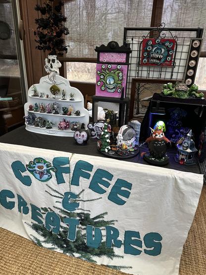 A test setup for a market booth on a porch. There is a weird white tree with black branches, a white display stand holding TTRPG minis, a beholder on  black and pink cabinet, a beholder cup rack, and many other very strange creatures.
From the front of the table, a fabric banner reading "coffee and creatures" is hanging. It is badly sewn on purpose, and the letters are turquoise against the white background.