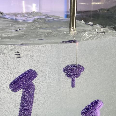 Close-up photo through the side of a large tank of clear gel. The surface of the gel is not flat, but contoured like hills, and the gel is filled with many tiny bubbles distributed throughout. A metal probe extends into the gel, printing a part that looks somewhat like a purple baby bottle nipple. Two completed purple parts that look like video game controllers are suspended to the left and further below within the gel.