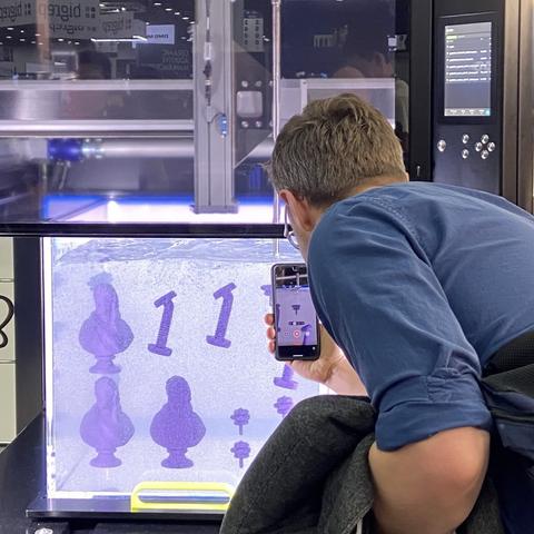 An expo visitor bends down in front of the vat of gel and printed parts, photographing the printer nozzle  with his phone. Purple silicone parts are suspended in the tank, and the gel is strongly illuminated in slightly blue light so it looks like they're floating in a glowing aquarium.