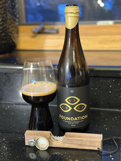 A stout glass of dark beer next to a tall brown bottle with a golden neck wax and a black label with three gold-rimmed orange eyes. In front is a wax cutter and bottle opener holding the bottle cap