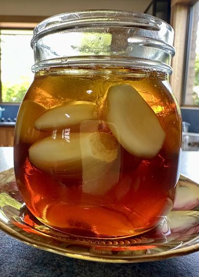 A glass jar filled with amber-colored honey, with peeled garlic cloves submerged in it. The jar is placed on a decorative plate and there is a blurred kitchen background.
