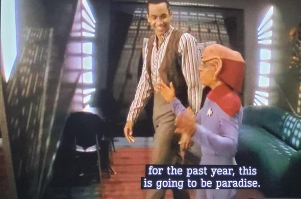 Jake Sisko smiling broadly in a striped shirt and open brown vest as he walks through the apartment with Nog. Nog is in his Starfleet Ensign uniform and gesticulates animatedly as he speaks, "for the past year, this is going to be paradise."
A green couch and black chair orange carpeting is shown.