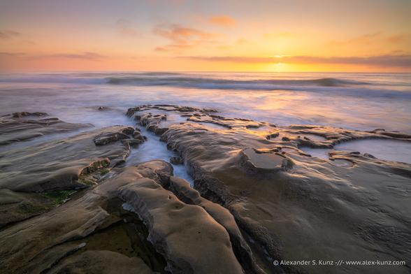 A seascape photo with tide pools and strange rock formations in the foreground, breaking waves rolling in, and the warm sun just above the horizon but behind a thin band of clouds.