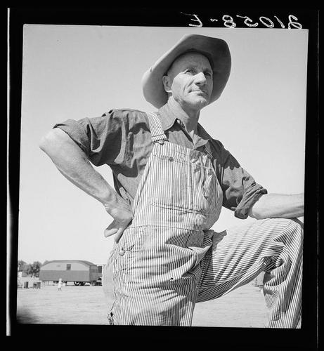 A black and white photograph of a man standing outdoors in a rural setting. He is wearing a wide-brimmed hat, overalls with vertical stripes, and has his hands on his hips. Behind him, there's an RV or trailer parked alongside the road. In the distance, another building can be seen through the sparse vegetation. The photo appears to have been taken during World War II era based on its style and quality. There is text overlay at the top of the image with a date "3-82016," suggesting it's either from that period or digitally altered for context.
