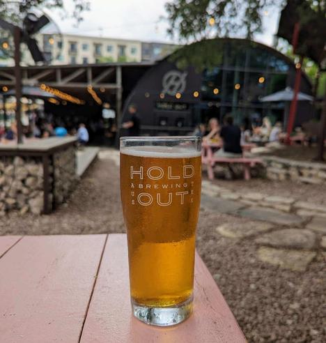 A pint glass of copper-colored beer sitting on a picnic table with the name "Hold Out Brewing" etched on it. In the background, a bit blurred out, is Hold Out Brewing itself, a brewery in Austin, Texas situated in a former Quonset hut. You can kind of see in the picture that there's a fair amount of outdoor, beer garden style space with gravel (instead of grass) and stonework. There are other people sitting at tables in the distance.