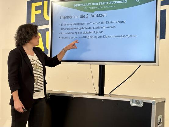 Prof. Dr. Claudia Reuter, 2. Vorsitzende des Digitalrats der Stadt Augsburg: Eine Frau mit dunklen, lockigen Haaren und schwarzem Blazer zeigt auf eine Präsentationsfolie. Titel: „Themen für die 2. Amtszeit“ des Digitalrats der Stadt Augsburg. Aufzählungspunkte zu Digitalisierungsthemen und Projektbegleitung.