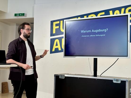 Benjamin Seuffert - Digitalsparte des Staatstheaters: Ein Mann mit dunkler Kleidung steht links neben einem großen Monitor. Die Präsentationsfolie zeigt die Frage „Warum Augsburg?“. Hintergrund: das gelbe „Future Week Augsburg“-Banner.