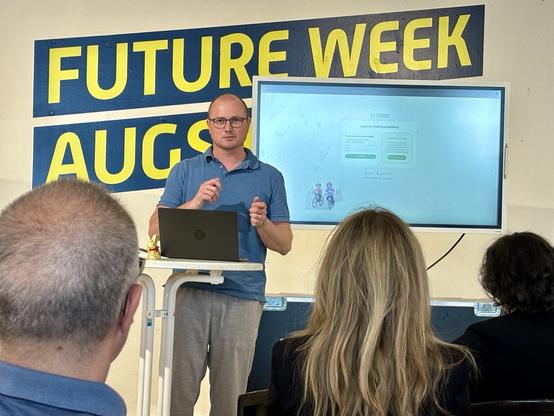 Dr. Thomas Eisenbarth: Ein Mann mit Brille und blauem Polohemd spricht vor Publikum. Er steht an einem Stehtisch mit Laptop. Hinter ihm ein großer Bildschirm. Im Hintergrund gelbes „Future Week Augsburg“-Banner an der Wand.