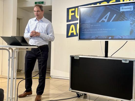 Prof. Dr. Ullrich Schilp - Uni Augsburg / Fraunhofer: Ein Mann im gestreiften Hemd hält einen Vortrag. Auf dem Bildschirm: „Wie KI und Generative KI die Produktion und Produktionsplanung revolutionieren“ – mit Fraunhofer IGCV-Logo. Er steht an einem Stehtisch mit Laptop, wirkt engagiert.
