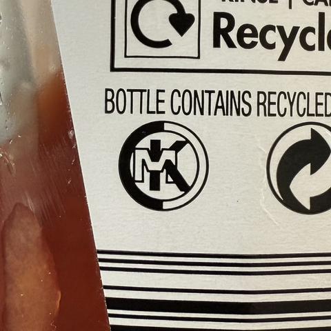 A close up of the label on a Heinz Tomato Ketchup plastic bottle (UK). Under the text “Bottle contains recycled plastic is the common plastics recycle logo and another logo consisting of;

- a circle with a thick outline. The left half is solid black and the right half is hollow with a thin black stroke
- inside the circle is a “MK” logotype. The M is hollow with a thin black stroke and the K is solid black.
