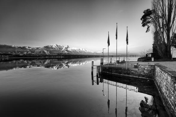 Schwarzweissfotografie eines ruhigen Sees mit Schiffsanlegestelle im Vorder- und Bergen im Hintergrund.