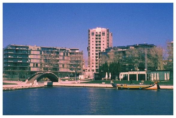 The cityscape of Aveiro in Portugal. In the foreground is the canal, with a tourist boat upon it. Behind are blocks of flats. The image is slightly colour shifted since Harman Phoenix film was decanted into Agfa Rapid film canisters. Taken with a 1940s Agfa Karat camera.