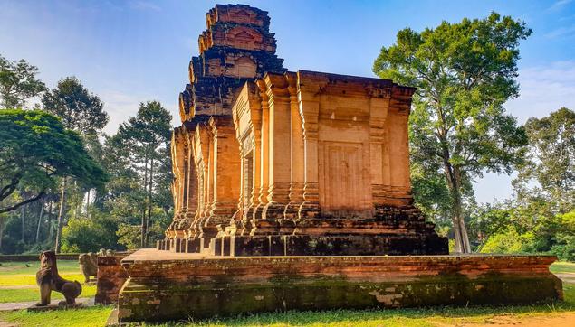 Photo shows an old temple, a row of five towers on a meter tall platform. Two of the towers still have a roof telling of an architectural style of early Angkor Empire. The side gates to the towers on the north side are sealed off. The gates to the east are open. The photo is taken slightly from the northeast corner. In front there are remains of lion guardian statues in various condition. It is an early morning just after sunrise and the temple is flooded by soft, warm sunlight. The temple is placed on green turf, with some red brown tracks and a stone pathway to leading up to the temple from the east/left. In the background there is a fundament for another building, presumably a library. Around the temple area the forest is forming a green border. The sky is light cloudy blue.