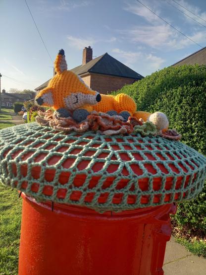 A knitted postbox topper on a red pillar box. It is an orange and white fox curled up asleep in some leaves and pine cones.
Edit: Actually, I think the pine cones are her feet!