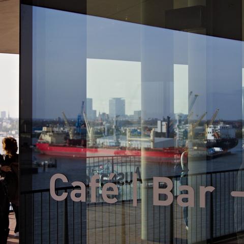 Blick auf der Elphi-Plaza durch eine verfensterte Ecke des Cafe.
Auf dem Fenster die Schrift "Cafe | Bar", im Fenster eine Spiegelung von Schiffen. Durchscheinend dann Hochhäuser.