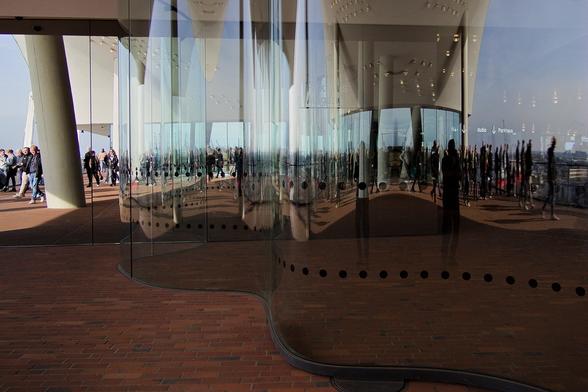 Blick von aussen auf die in wellenform geschwungene Glasfront der Elphi-Plaza. In dem Glas spiegeln sich verzerrt wie im einem Zerrspiegel die Besucher der Plaza. Links ist noch ein Teil der umführenden Terrasse mit Menschen zu sehen. Durch die Glasspiegelungen scheinen Leuchtschriften und Belöeuchtung von innen durch.