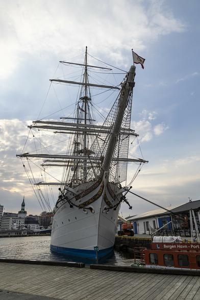 A portrait view of a three-masted sailing ship viewed from the dockside where it is located and looking at it almost directly from the front. It appears narrow and tall and almost leaning slightly to the right because we're just off the centre and the bowsprit reaches up and over us just to the right. The masts have numerous yard arms on them but all the sails are furled. It's early evening here and there are some low buildings to the right on the dock and across the water on the left more buildings of this city are seen in shadow. The sky has patchy cloud but with the sun in the process of setting somewhere hidden from view there's a fairly cool, subdued feel.