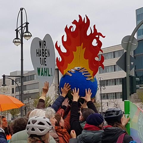 Menschen halten ein Modell der brennenden Erde in die Luft. Daneben ein "Wir haben die Wahl"-Plakat.