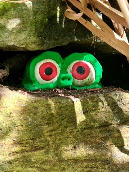 A sculpture of a small green creature with big red eyes looking out from a crevice between two stones in an old wall.