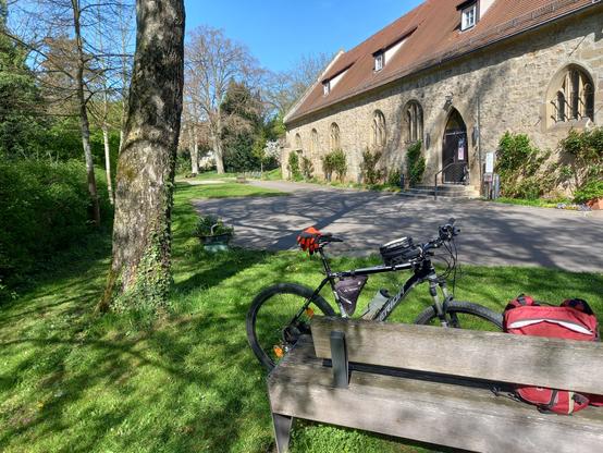 Rechts eine Parkbank im Gras, an der ein Fahrrad und ein Rucksack lehnen. Links der Stamm eines Baums. Im Hintergrund ein altes Gebäude (das Hölderlinhaus in Lauffen/Neckar)